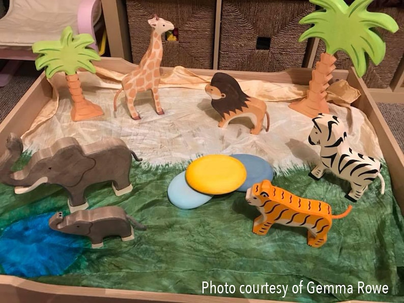 May include: A wooden play set with a variety of animal figurines, including a giraffe, zebra, tiger, elephant, and lion. The set also includes two palm trees and a green and brown play mat.