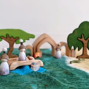 May include: Two wooden figurines with brown hats and white faces are on a wooden bridge over a blue river. The bridge is made of wood and has a natural finish. The river is surrounded by green grass and trees. There are two wooden trees in the background, one with a green canopy and the other with a brown trunk and green leaves.