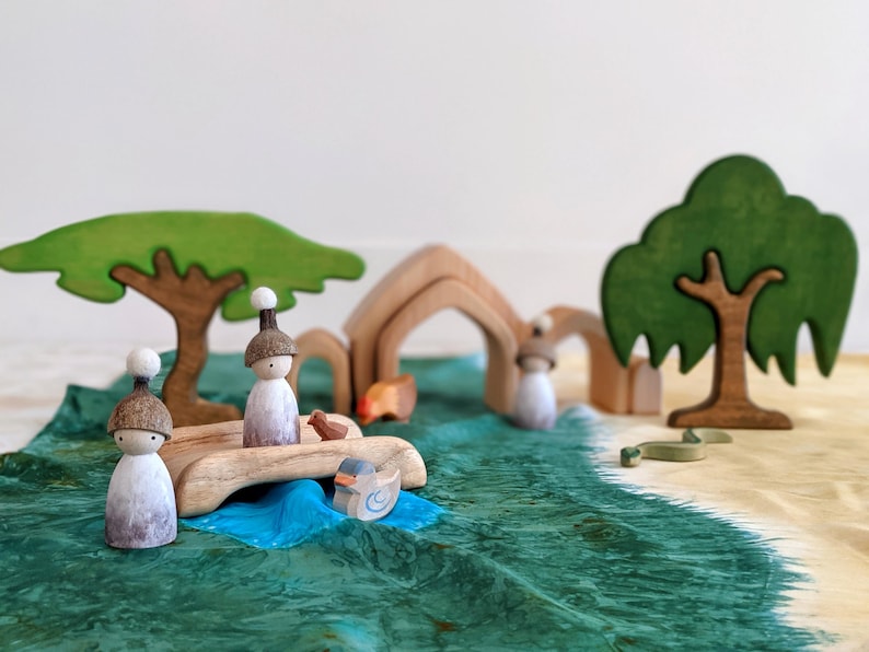 May include: Two wooden figurines with brown hats and white faces are on a wooden bridge over a blue river. The bridge is made of wood and has a natural finish. The river is surrounded by green grass and trees. There are two wooden trees in the background, one with a green canopy and the other with a brown trunk and green leaves.
