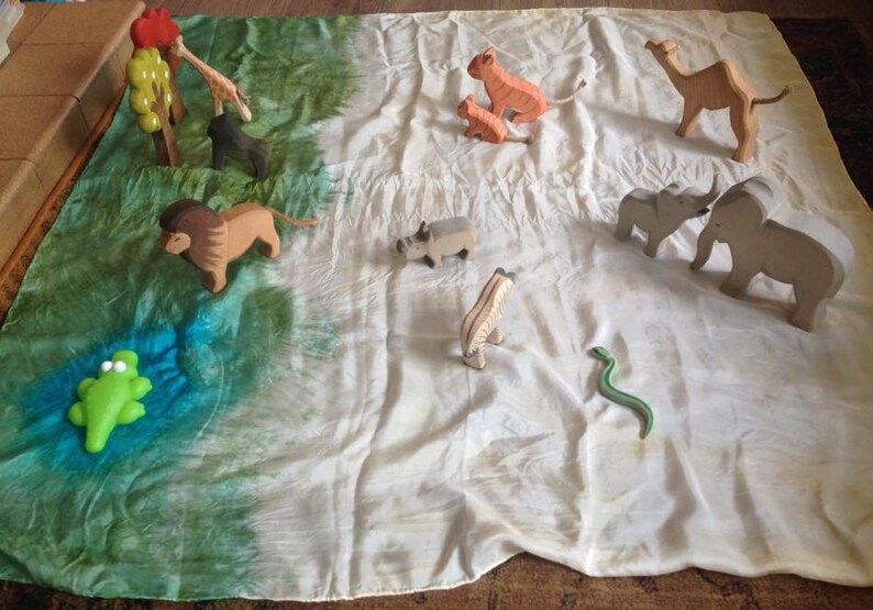May include: A set of wooden animal figurines, including a lion, zebra, giraffe, camel, elephant, rhino, snake, and frog, arranged on a green and white fabric play mat.