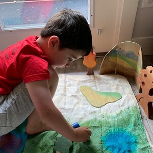 May include: A child plays with wooden toy trees and a blue paint roller on a green and blue fabric play mat.