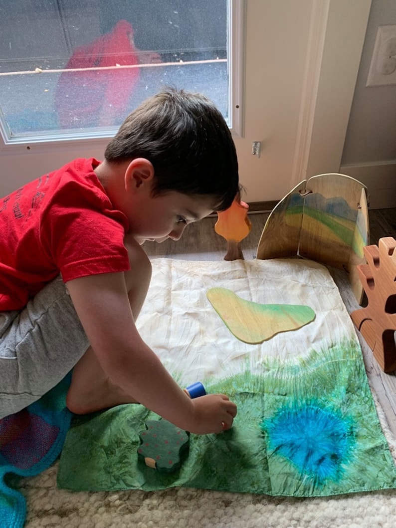 May include: A child plays with wooden toy trees and a blue paint roller on a green and blue fabric play mat.