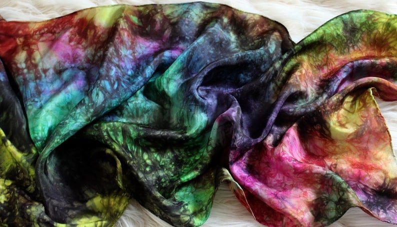 May include: A colorful silk scarf with a tie-dye pattern in shades of green, blue, purple, pink, and yellow. The scarf is draped over a white surface.
