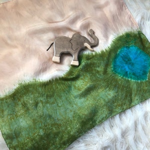 May include: A silk scarf with a tie-dye pattern in shades of green, brown, and blue. A wooden elephant toy is placed on the scarf.