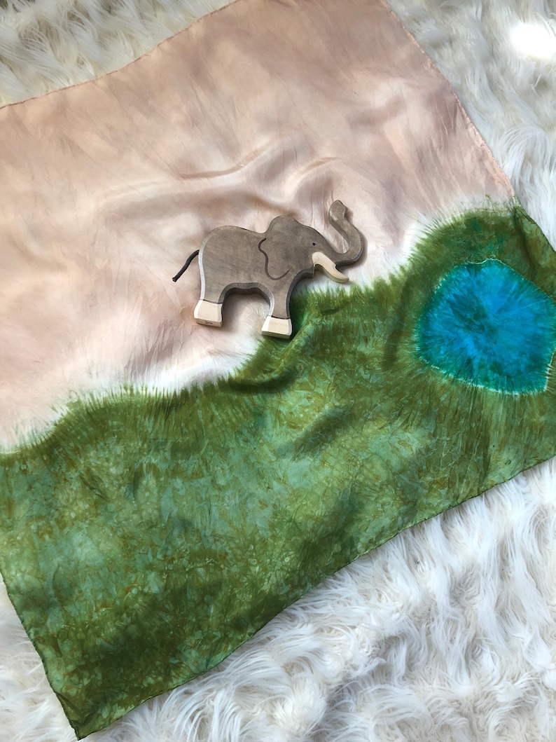 May include: A silk scarf with a tie-dye pattern in shades of green, brown, and blue. A wooden elephant toy is placed on the scarf.
