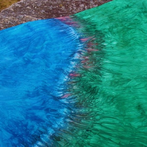 Op de afbeelding: Een levendig textiel met een aquareleffect, met een kleurengradiënt. De stof gaat over van een diepblauw aan de linkerkant naar een heldergroen aan de rechterkant, met een band van roze en rood in het midden. De textuur lijkt zacht en vloeiend.