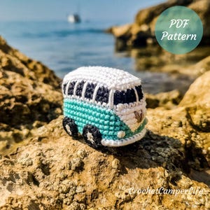 May include: A crocheted miniature camper van in white, teal, and black, with black wheels and windows. The van is set against a blurred background of rocks, sea, and sky. The text "PDF Pattern" is in a green circle.