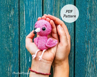 May include: A handmade pink crochet flamingo with black eyes, a white beak, and white legs is held in cupped hands. The flamingo is set against a teal wooden background. A white circle with the text "PDF Pattern" is in the upper right corner.