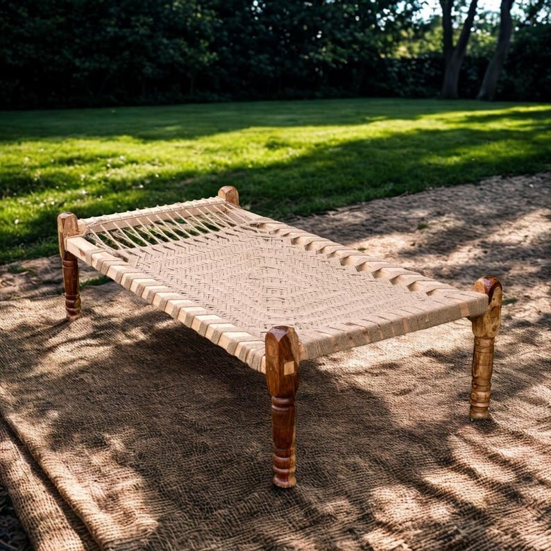 May include: A woven rope bed with a rectangular frame and four wooden legs. The bed's surface is made of tightly woven natural-colored rope, and the legs are a dark brown color. The bed is outdoors on a brown surface, with green grass and trees in the background.
