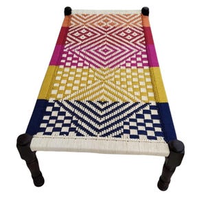 May include: A woven bed frame with a colorful geometric pattern. The bed features a rectangular design with dark wooden legs and a woven surface in shades of red, pink, yellow, and blue.