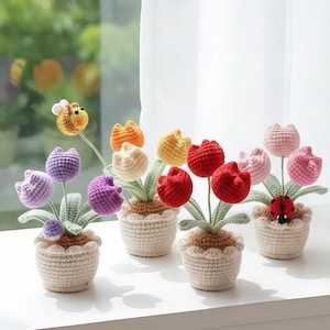 Può includere: Una collezione di tulipani all'uncinetto fatti a mano in piccoli vasi. I tulipani sono in vari colori, tra cui viola, arancione, giallo e rosso. Sono inclusi anche un'ape e una coccinella all'uncinetto. I vasi sono beige.