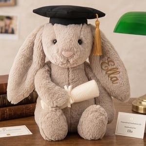 Peut inclure: Un lapin en peluche gris clair portant une toque de graduation noire avec un gland doré. Le lapin tient un diplôme enroulé et attaché avec un ruban blanc. Le nom "Ella" est brodé sur l'oreille. Un cadeau de remise de diplôme.