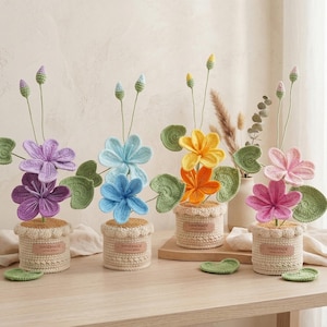 May include: A collection of handmade crochet flowers in various colors, including purple, blue, yellow, orange, and pink. Each flower is potted in a small, beige crocheted pot with a "HANDMADE" label. Green leaves and buds accompany the flowers.