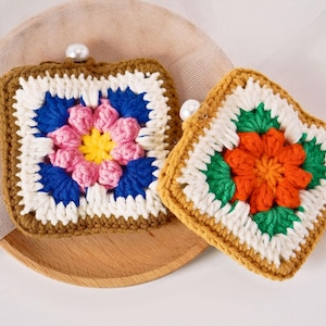 Może przedstawiać: Dwa ręcznie robione podkładki z szydełka z kwiatowymi wzorami. Jedna podkładka ma różowy, żółty i niebieski kwiat, a druga pomarańczowy i zielony. Obie są kwadratowe z perłowym akcentem.