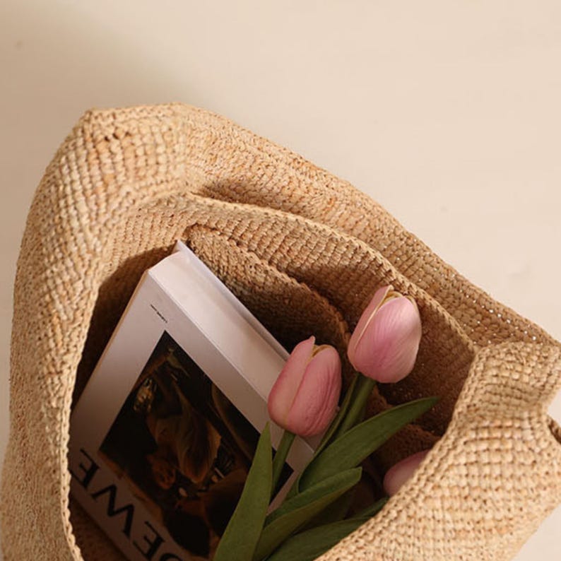 May include: A woven straw tote bag with a book and pink tulips inside. The bag is a light tan color and has a textured appearance. The book has a white cover and the flowers have green stems and leaves.
