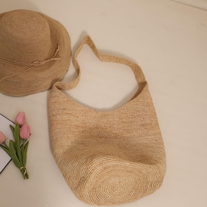 May include: A natural-toned woven straw shoulder bag with a matching wide-brimmed hat. The bag has a curved top and a long strap. The hat has a rounded crown and a tied band. The items are made of a light brown straw material.