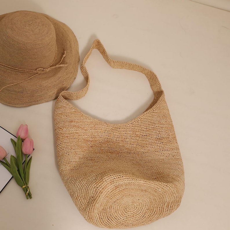 May include: A natural-toned woven straw shoulder bag with a matching wide-brimmed hat. The bag has a curved top and a long strap. The hat has a rounded crown and a tied band. The items are made of a light brown straw material.