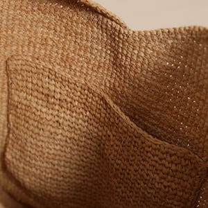 May include: Close-up of a woven bag made from natural fibers. The bag has a textured surface with a pocket detail. The color is a warm, earthy brown, suggesting a handmade or artisanal product. The weave is tight, indicating durability.