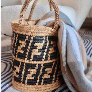 May include: A cylindrical woven straw tote bag with a black geometric design. The bag features braided handles and a natural colour. A folded blanket in blue, tan, and white is draped over the side.