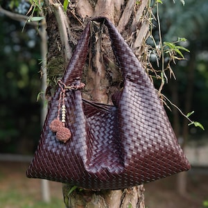 May include: A large, dark brown woven leather tote bag with a triangular shape. The bag hangs from a tree branch and features a drawstring closure with decorative tassels. The bag's texture and color suggest a handcrafted design.