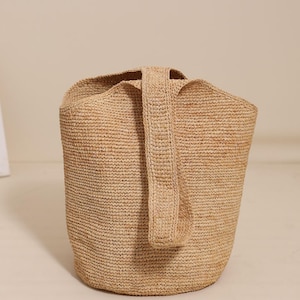May include: A woven raffia tote bag in a natural beige color. The bag has a cylindrical shape with a single, sturdy handle. The texture of the raffia is visible, adding to the bag's rustic charm. The bag is suitable for carrying various items.