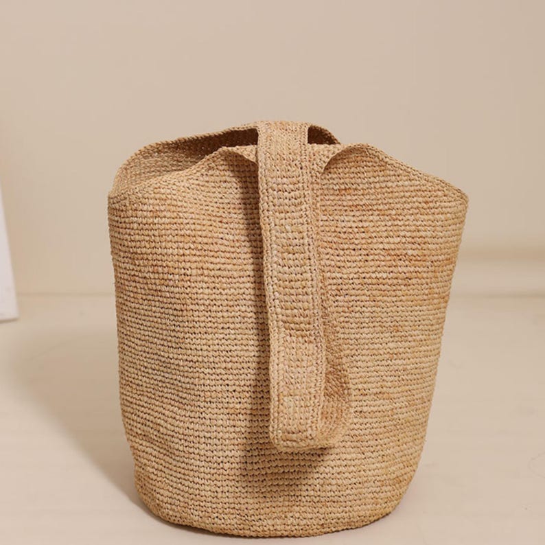 May include: A woven raffia tote bag in a natural beige color. The bag has a cylindrical shape with a single, sturdy handle. The texture of the raffia is visible, adding to the bag's rustic charm. The bag is suitable for carrying various items.