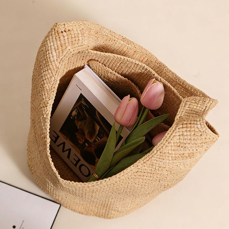 May include: A woven straw tote bag with a natural beige color. Inside the bag is a book with the word "LOEWE" visible, and a bouquet of pink tulips. The bag is open, revealing its contents.