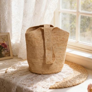 May include: A beige woven tote bag with a single handle. The bag is cylindrical with a wide opening and a rectangular handle. The bag is sitting on a white lace cloth and a round woven mat.