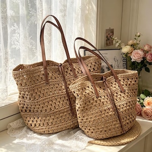 May include: Two tan woven straw tote bags with brown leather handles and drawstring closures. The bags feature an open crochet pattern and a structured base. The larger bag is positioned behind the smaller bag, both displaying a natural texture.