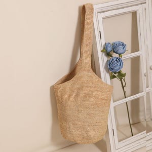 May include: A woven beige tote bag with a single handle. The bag has a bucket shape and is hanging against a white wall with a window frame and blue flowers.