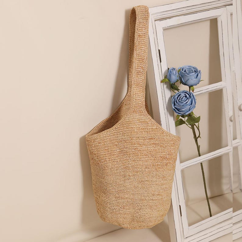 May include: A woven beige tote bag with a single handle. The bag has a bucket shape and is hanging against a white wall with a window frame and blue flowers.