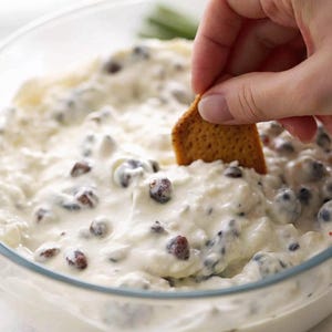 May include: A clear glass bowl filled with a creamy, white dip speckled with dark, round ingredients. A hand dips a square cracker into the dip. The dip appears to be a savory appetizer.