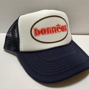 Può includere: Cappellino da camionista blu navy e bianco con la scritta "Bonneau" in rosso all'interno di un ovale marrone. Il cappello ha un retro in rete e una visiera curva.