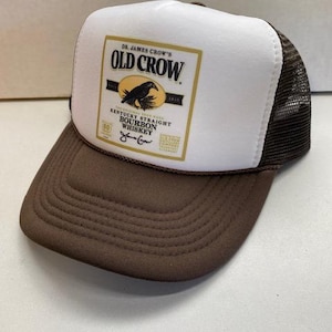 May include: A brown and white trucker hat with a brown peak and mesh back. The front panel displays a label with the text "Dr. James Crow's Old Crow Kentucky Straight Bourbon Whiskey" and a crow illustration.