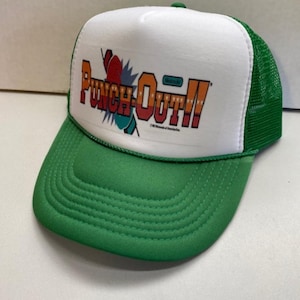 Peut inclure: Casquette de camionneur verte et blanche avec le texte "Punch-Out!!" en orange, rouge et bleu. La casquette a une visière verte et un dos en filet, avec un panneau avant blanc affichant le logo du jeu.