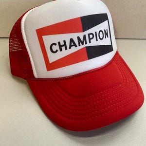 Puede incluir: Gorra de camionero roja y blanca con el logotipo de Champion. El logotipo es un rectángulo con la palabra "CHAMPION" en negro. La gorra tiene una visera roja y una parte trasera de malla.