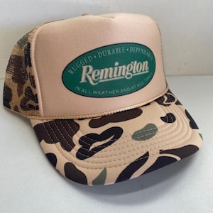 Puede incluir: Gorra de camionero de color canela y camuflaje con el logotipo de Remington en un óvalo verde. La gorra presenta una parte trasera de malla, una visera curva y el texto "RUGGED DURABLE DEPENDABLE IN ALL WEATHER AND AT ALL TIME".
