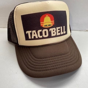 May include: A brown and tan trucker hat featuring the Taco Bell logo. The logo is a red bell with a yellow outline, set against a dark brown rectangle. The text "TACO BELL" is printed in white.