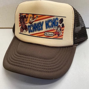 Op de afbeelding: Bruine en beige truckerpet met het Donkey Kong-logo en -personages. De pet heeft een bruine klep en mesh achterkant, met het Nintendo-logo op de voorkant.