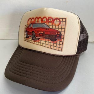 May include: A brown and tan trucker hat featuring a red Camaro car graphic. The word "CAMARO" is printed above the car in orange letters. The hat has a brown brim and a mesh back.