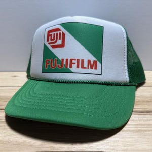 Peut inclure: Casquette de camionneur Fujifilm verte et blanche. Le panneau avant présente le logo Fujifilm en rouge et blanc, sur un fond vert et blanc. La visière et le dos en filet sont verts.
