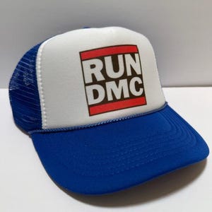 May include: A blue and white trucker hat with the text "RUN DMC" in a red and white rectangular design on the front. The hat has a blue brim and a white front panel with a mesh back.