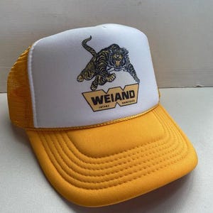 以下が含まれることがあります： トラのグラフィックと「WEIAND INTAKE MANIFOLDS」の文字が入った、黄色と白のトラッカーハット。帽子は黄色のメッシュバックと黄色のつばが付いています。