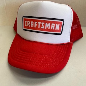 May include: A red and white trucker hat with the word "CRAFTSMAN" in a red rectangle on the front. The hat has a red brim and mesh backing.