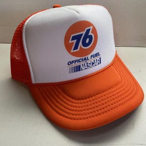 May include: Orange and white trucker hat with the number 76 in a circle on the front. The hat has a mesh back and a curved brim. The text "OFFICIAL FUEL NASCAR" is printed below the number.