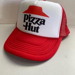Puede incluir: Gorra de camionero roja y blanca de Pizza Hut. La gorra tiene una visera roja y una parte trasera de malla, con el logotipo de Pizza Hut en negro y rojo en el panel frontal. Un estilo de gorra clásico.