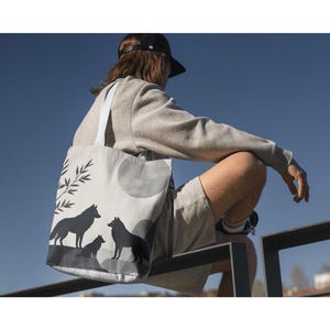 May include: A white tote bag featuring a black silhouette of wolves under a full moon and bamboo branches. The bag has white straps and is carried by a person wearing a gray sweatshirt and shorts.