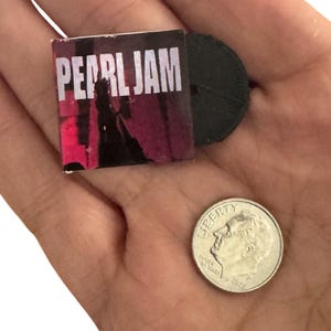 May include: Miniature replica of a Pearl Jam album cover with the band's name in white block letters on a pink and black background. A small black vinyl record is next to the album. A dime is also in the image.