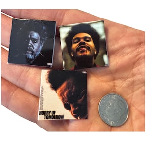 May include: Three small square images featuring portraits of a musician, and one coin. One image has the text "HURRY UP TOMORROW". The images are held in a hand.