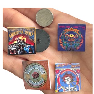 May include: Miniature Grateful Dead album covers and a vinyl record. The album covers feature colorful artwork and the band's name. A silver coin is also present. The items are small and detailed, perfect for collectors or craft projects.
